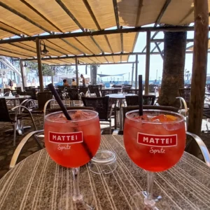 Zwei Gläser mit orangerotem Getränk und Aufklebern 'MATTEI Spritz' auf einem Tisch unter einem Sonnenschutz im Freien mit Blick auf Meer