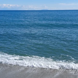Sanft anbrandende Wellen am Strand unter blauem Himmel mit vereinzelten Wolken, weiter Horizont über dem Meer bei Korsika.