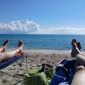 Zwei ausgestreckte Beine auf Liegestühlen am Sandstrand mit Blick auf das Meer und bewölkten Himmel auf Korsika.