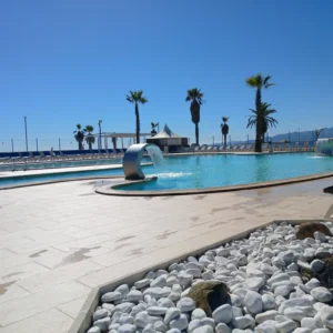 Pool mit Wasserfallfontäne, umgeben von Palmen und Liegestühlen unter klarem blauem Himmel auf Korsika.