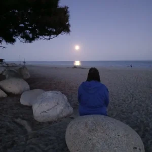 Eine Person in einer blauen Kapuzenjacke sitzt von hinten gesehen auf einem großen, runden Stein an einem Sandstrand und blickt in der Abenddämmerung auf das Meer, während sich der helle Vollmond auf der Wasseroberfläche spiegelt.