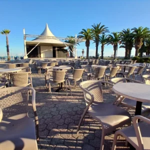 Leere Terrasse mit vielen runden Tischen und Stühlen aus Kunststoff, im Hintergrund eine Bühne mit weißem Zeltdach und Palmen, sonniger Himmel.