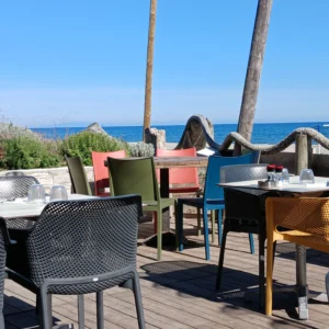 Mehrfarbige Kunststoffstühle und gedeckte Tische auf Holzterrasse mit Blick auf das Meer und blauen Himmel.