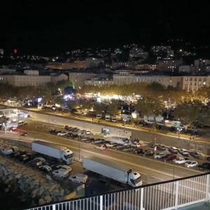 Eine nächtliche Aufnahme aus erhöhter Perspektive auf einen beleuchteten Hafenbereich und eine Küstenstadt in Korsika, bei der im Vordergrund parkende Lkw und Pkw an einer Uferpromenade stehen und im Hintergrund ein von Bäumen gesäumter, heller Platz sowie Wohnhäuser an einem Berghang zu sehen sind.
