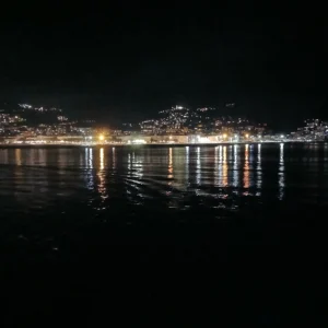 Nächtliche Panoramaaufnahme von Bastia auf Korsika mit beleuchteter Küstenlinie und Lichtern auf den Hügeln, reflektiert im Wasser.