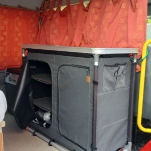 Busumbau Der Innenraum eines roten Ford Transit Camping-Vans mit einem grauen Falt-Campingküchenschrank, einer Kühlbox von Berger und orangefarbenen Vorhängen.