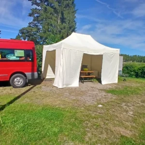 Ein roter Kastenwagen parkt auf einer Wiese neben einem großen, weißen Faltzelt mit Seitenwänden, in dem ein Holztisch steht.