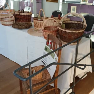 Verschiedene handgeflochtene Weidenkörbe stehen aufgereiht auf einem langen, mit einer weißen Decke verkleideten Tisch in einem Ausstellungsraum mit Holzboden.