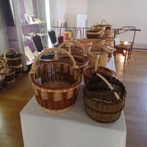 Handgeflochtene Weidenkörbe in verschiedenen Größen und Brauntönen stehen auf einem langen, weiß gedeckten Tisch in einem hellen Ausstellungsraum mit Parkettboden.