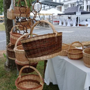 Handgeflochtene Weidenkörbe der Korbmacherei Kreative Finger, die an dekorativen Weidenketten von einem Marktzeit herabhängen, im Hintergrund ein gepflasterter Platz mit Gebäuden.
