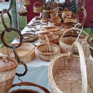 Bauernmarkt 7/2026 Ein reich bestückter Marktstand der Korbmacherei Kreative Finger im Freien, fotografiert durch dekorativ aufgehängte Weidenringe im Vordergrund, mit zahlreichen geflochtenen Körben auf einem langen Tisch.