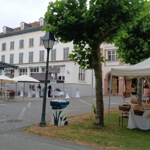 Marktstand vor dem Kurhaushotel Ein weißes Marktzeit der Korbmacherei Kreative Finger mit geflochtenen Weidenkörben auf einem Platz vor einem großen, historischen Gebäude mit Außengastronomie.