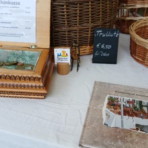 Bauernmarkt 7/2026 Ein Verkaufsstand der Korbmacherei Kreative Finger mit einer hölzernen Schönkasse voller Münzen, geflochtenen Weidenkörben und einem Fotobuch auf einer weißen Tischdecke.