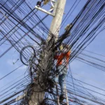 Ein Arbeiter in einer orangefarbenen Warnweste klettert auf einer Holzleiter an einem Betonstrommast empor, der von einem extremen, chaotischen Gewirr aus unzähligen Strom- und Telekommunikationskabeln vor einem blauen Himmel umgeben ist.