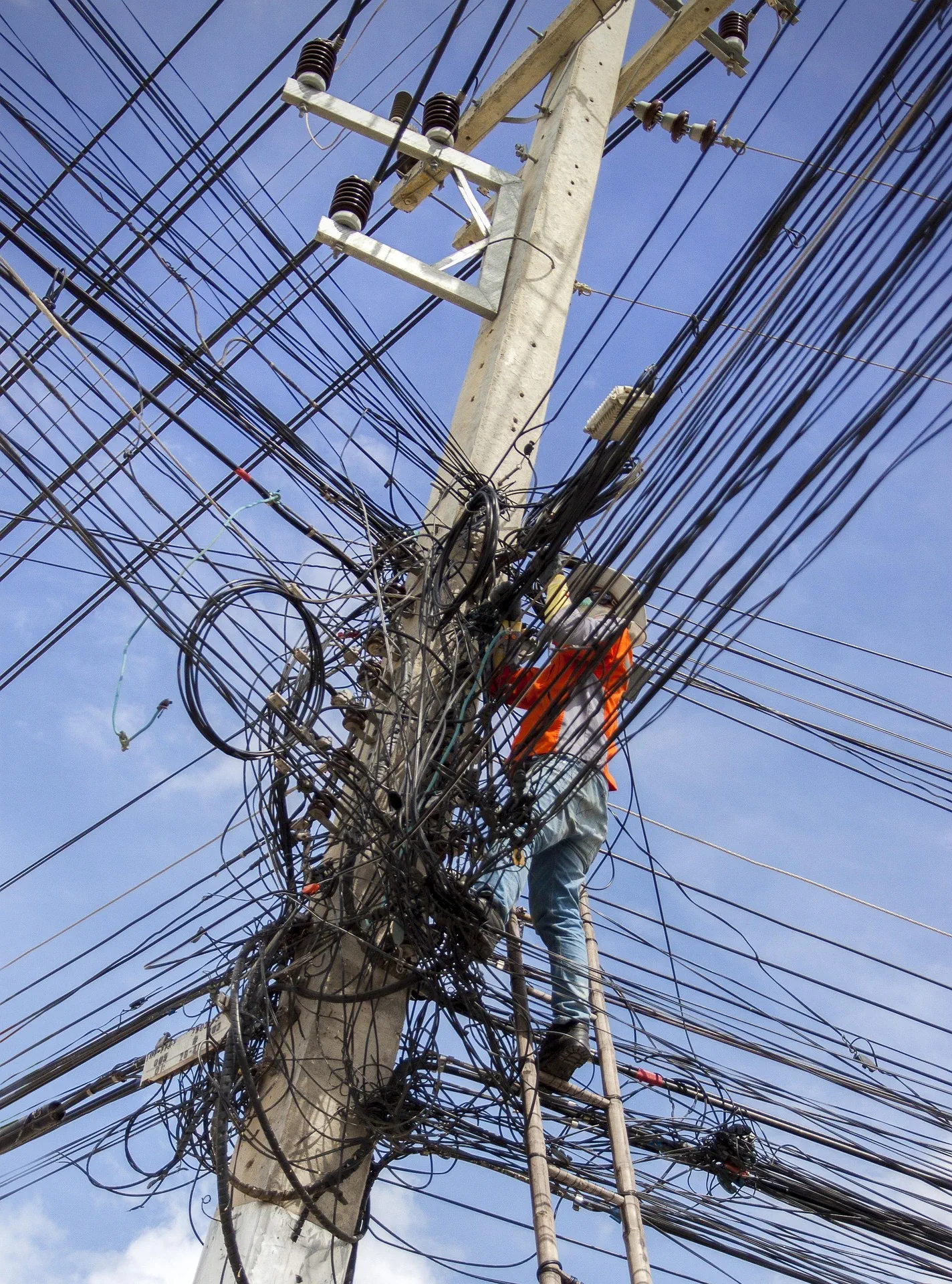 Ein Arbeiter in einer orangefarbenen Warnweste klettert auf einer Holzleiter an einem Betonstrommast empor, der von einem extremen, chaotischen Gewirr aus unzähligen Strom- und Telekommunikationskabeln vor einem blauen Himmel umgeben ist.